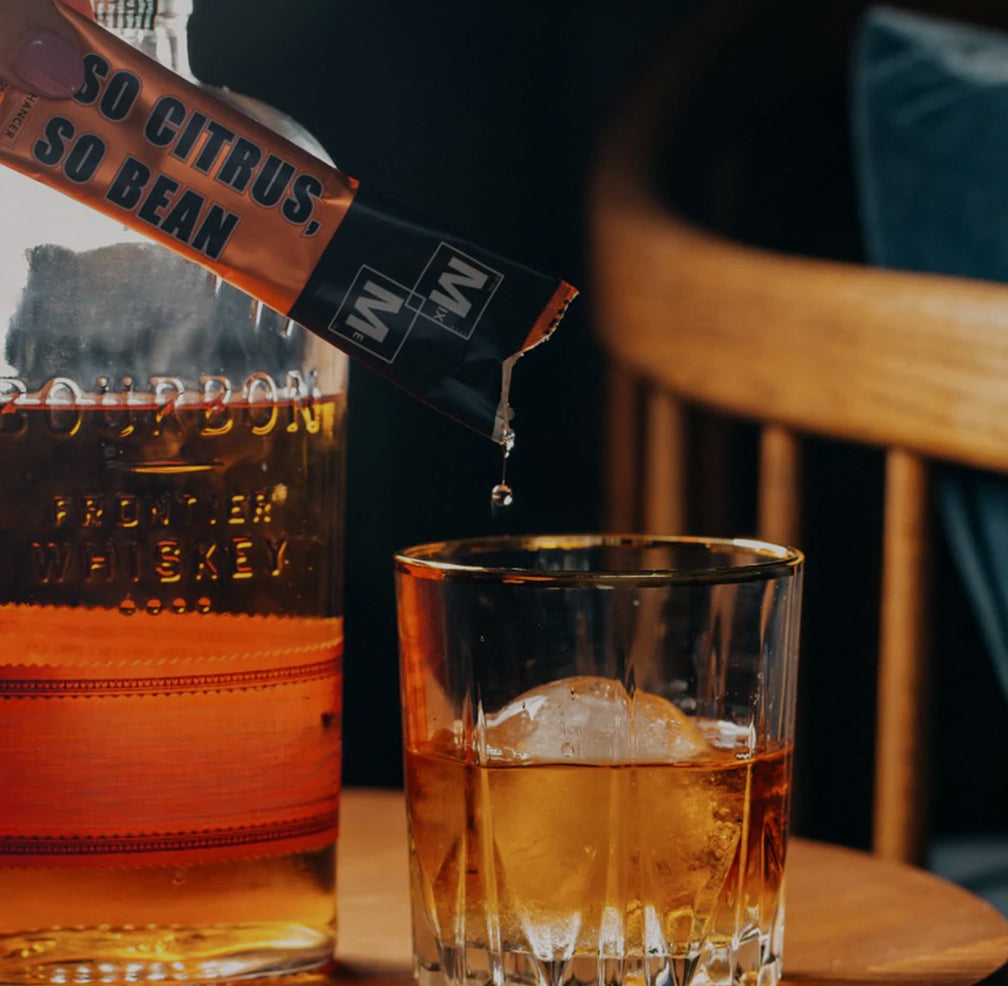 Bourbon bottle with a glass of whiskey and a packet labeled 'So Citrus, So Bean' on a wooden surfac