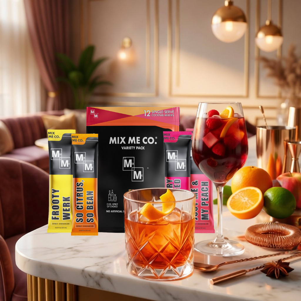 Table with Mix Me Co. drink mix packets, glasses of cocktails, and fruits in a cozy indoor setting.